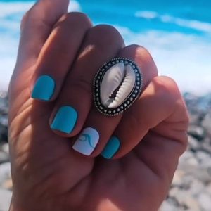 Primer plano de una mano con manicura turquesa luciendo un anillo regulable de zamak con una concha natural cauri frente a una playa de piedras y mar azul.