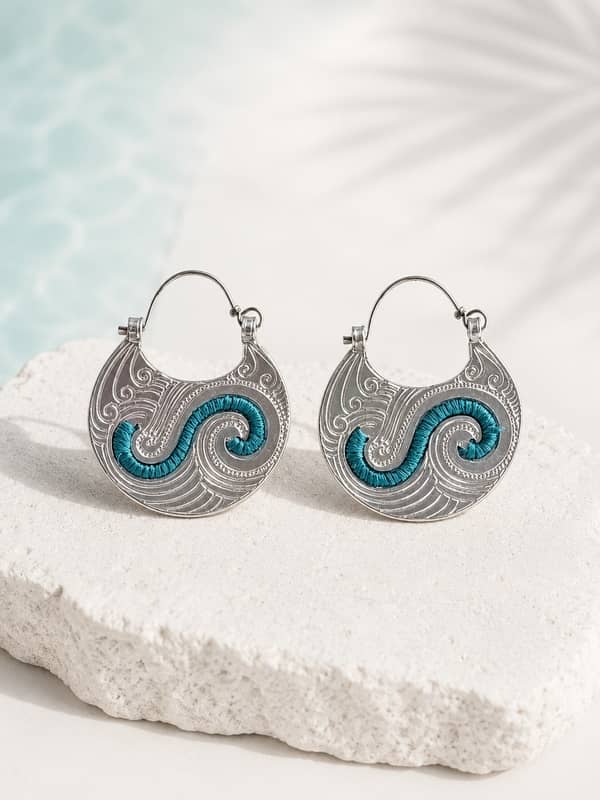 Par de pendientes de argollas canarias artesanales de aluminio grabado con una S central tejida en hilo encerado turquesa, apoyados sobre una piedra blanca con fondo playero.