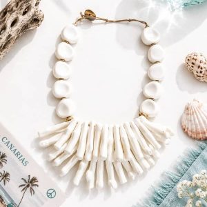 Collar de cuentas y ramificaciones de coral de resina en color blanco mate con cordón ajustable rústico, fotografiado en ambiente costero junto a conchas marinas y madera flotante.