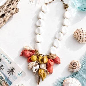 Collar de cuentas grandes de resina blanca con un colgante central de caracolas en tonos teja, ocre y blanco, colocado sobre un fondo playero decorado con conchas y una postal de Canarias.