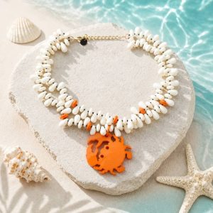 Collar doble de cuentas de resina blancas y detalles coral sobre una roca de playa, con un colgante central de cangrejo naranja brillante, rodeado de conchas y una estrella de mar.