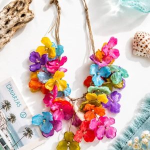 Collar de flores tridimensionales de resina en colores brillantes fucsia, amarillo, azul y naranja, con cordón ajustable de estilo natural, colocado sobre un fondo playero decorado con madera flotante y conchas marinas.