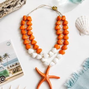 Collar de doble vuelta hecho con cuentas de resina naranjas y blancas en forma de conchas y un colgante central de estrella de mar naranja, colocado sobre un fondo veraniego con caracolas y una postal de playa.