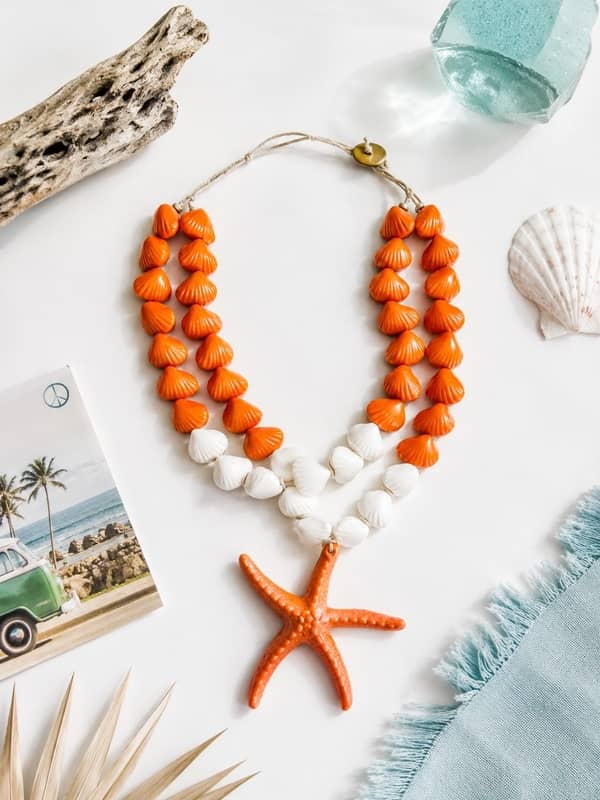 Collar de doble vuelta hecho con cuentas de resina naranjas y blancas en forma de conchas y un colgante central de estrella de mar naranja, colocado sobre un fondo veraniego con caracolas y una postal de playa.