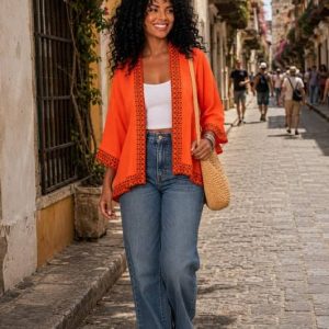 Mujer sonriente con cabello rizado camina por una calle colonial vistiendo un kimono corto naranja con bordados étnicos, combinado con vaqueros y top blanco.