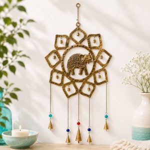 Móvil decorativo de pared de cobre dorado con forma de mandala de estrella y un elefante labrado en el centro, con campanas colgantes y cuentas de colores, colgado en una pared blanca junto a una vela encendida y flores.