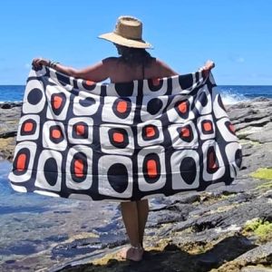 Mujer de espaldas con sombrero de paja sosteniendo al aire un pareo de viscosa con estampado geométrico retro en negro, blanco y naranja frente al mar.