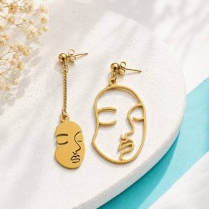 areja de pendientes asimétricos de acero dorado con formas de rostros artísticos, uno corto y calado, y el otro colgante con cadena fina sobre una superficie blanca.