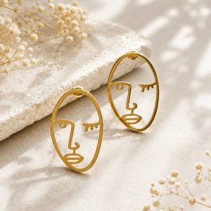 Pendientes de tuerca en acero dorado con forma de silueta de rostro abstracto calado con un ojo guiñado, sobre un fondo de piedra texturizada.
