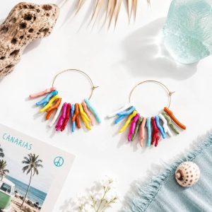Par de pendientes de aro dorados con cuentas colgantes que imitan ramitas de coral de resina multicolor, colocados en una composición playera junto a una postal de Canarias y una caracola.