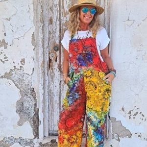 Peto largo de mujer de estilo boho chic con estampado tie-dye multicolor y dibujos de soles celestiales, combinado con una camiseta blanca, sombrero de paja y gafas de sol, posando frente a una puerta rústica de madera antigua.