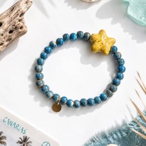 Pulsera elástica de cuentas de cerámica en color azul océano con un dije central en forma de estrella de mar de color amarillo, colocada sobre un fondo playero con caracolas marinas.