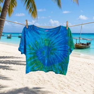 Camiseta corta tipo crop top de mujer con estampado tie-dye artesanal en tonos azul y verde, colgada con pinzas de madera en una cuerda de ropa sobre una playa de arena blanca y mar turquesa.