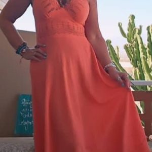 Mujer sonriente posando con vestido largo de color naranja coral de estilo boho chic, confeccionado en algodón 100% con escote halter y cuerpo bordado.