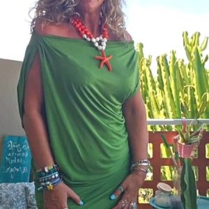 Mujer sonriente en una terraza vistiendo un vestido corto drapeado de color verde con hombros descubiertos, combinado con un collar llamativo de estrella de mar naranja y blanca.