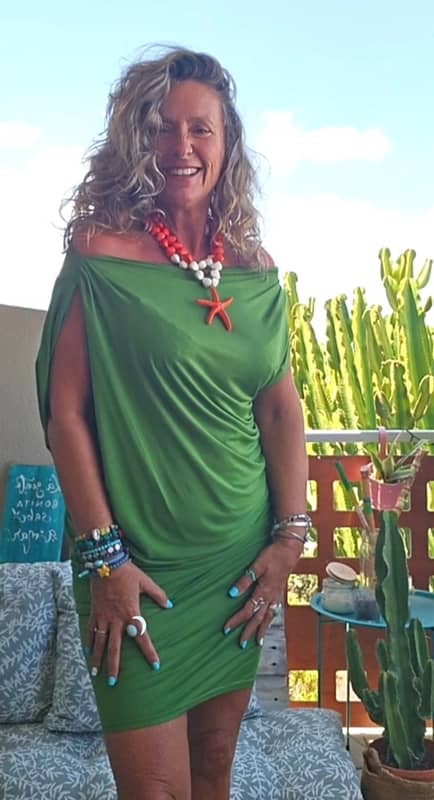 Mujer sonriente en una terraza vistiendo un vestido corto drapeado de color verde con hombros descubiertos, combinado con un collar llamativo de estrella de mar naranja y blanca.