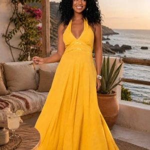 Mujer sonriente con pelo rizado posa en una terraza junto al mar vistiendo un vestido largo amarillo de algodón con escote halter para atar al cuello.