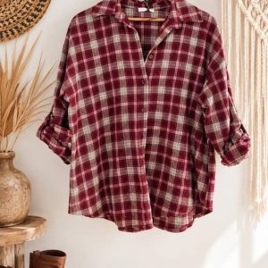 Camisa de cuadros de mujer en tonos granate y beige de estilo boho chic colgada en un perchero de madera rústica al lado de un sombrero de paja y unas botas camperas marrones de la tienda Sawabona.