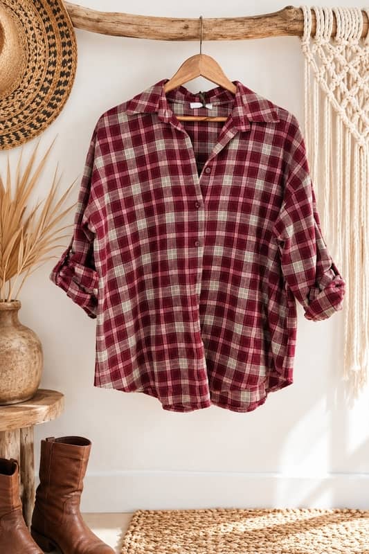 Camisa de cuadros de mujer en tonos granate y beige de estilo boho chic colgada en un perchero de madera rústica al lado de un sombrero de paja y unas botas camperas marrones de la tienda Sawabona.