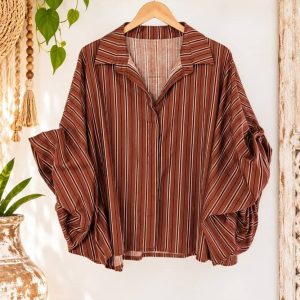 Camisa de rayas verticales color marrón terracota con mangas abullonadas fruncidas de Sawabona colgada en perchero de madera rústica.