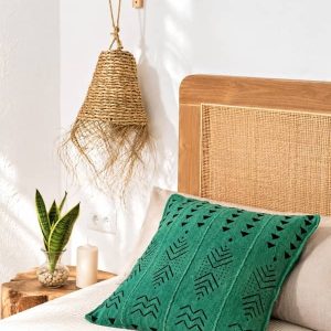 Cojín de bogolán africano verde de la tienda Sawabona decorando una cama con cabecero de ratán junto a una lámpara de esparto y una planta.