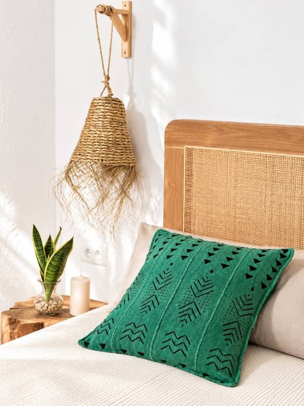 Cojín de bogolán africano verde de la tienda Sawabona decorando una cama con cabecero de ratán junto a una lámpara de esparto y una planta.