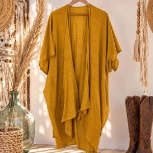 Kimono de la India color mostaza de algodón de Sawabona colgado de una percha en un perchero de rama rústica, decorado con damajuana de cristal, pampas secas y unas botas camperas marrones a los pies.
