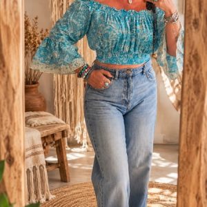 Mujer sonriendo frente al espejo vistiendo top corto boho azul turquesa con estampado paisley, hombros descubiertos y pantalón vaquero claro de talle alto.
